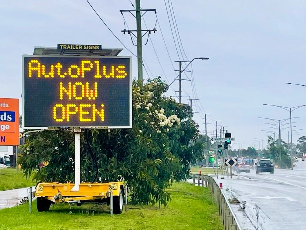 Bright amber VMS trailer sign for AutoPlus Autoexperts advertising “AutoPlus now open” near outlet in Cranbourne West
